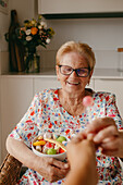 Eine fröhliche ältere Frau mit Brille lächelt, als ihr in einer gemütlichen Küche eine Schüssel mit frischem Obstsalat gereicht wird