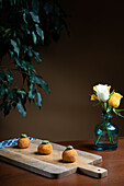 Drei goldene Arancini-Bällchen aus Reis und Käse, garniert mit Kräutern, präsentiert auf einem rustikalen Holzbrett, mit einer Vase mit gelben Rosen im Hintergrund.