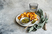 Golden roasted potatoes on a white plate with green pesto in a small bowl, a glass of water, and cutlery wrapped in a green napkin on a textured grey background