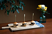 Three gourmet mini beef burgers topped with melted cheddar cheese, served on a rustic wooden board beside a vase of fresh yellow roses.