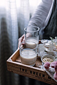 Zwei klare Gläser mit frischer, hausgemachter Hafermilch, präsentiert auf einem Holztablett mit rohem Hafer und Blumen, die einen gesunden Lebensstil unterstreichen