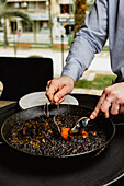 Eine Person serviert eine Portion traditioneller spanischer schwarzer Paella, die in einer großen, flachen Pfanne gekocht wird, wobei die appetitlichen Meeresfrüchte hervorgehoben werden