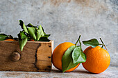 Reife Orangen mit frischen grünen Blättern werden ausgestellt, einige liegen auf einer strukturierten Oberfläche, andere sind in einer Holzkiste eingebettet und vermitteln den Eindruck einer frischen Ernte