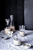 A serene tabletop scene featuring homemade oat milk in glass bottles and a tumbler, accented with raw oats and orchid flowers