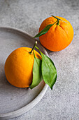 Nahaufnahme von zwei reifen Orangen mit Stängeln und einem grünen Blatt auf einem strukturierten grauen Teller, mit einem Weichzeichner-Hintergrund
