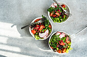 Top view of three bowls of healthy vegetable salad with lettuce, arugula, radishes, cherry tomatoes, and pomegranate seeds on a concrete background