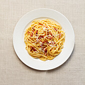 A top view of a classic Italian spaghetti carbonara, garnished with grated cheese and crispy bacon, served on a white textured background.
