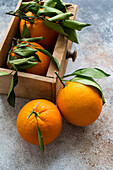 Top view of ripe oranges with fresh green leaves attached are displayed, some resting on a textured surface and others nestled in a wooden crate, evoking a sense of fresh harvest