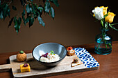 A variety of sophisticated canapés displayed on a wooden serving board, complemented by a bowl of creamy risotto and a vase of fresh yellow roses.