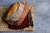 A crusty loaf of homemade rye sourdough bread placed on a woven mat with a dark red napkin and a bread knife