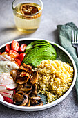 Close-up of a balanced lunch with a portion of bulgur wheat, a fried egg, avocado slices, cherry tomatoes, radishes, and brown mushrooms, accompanied by a glass of lemon water