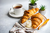 A breakfast setup with two golden croissants on a plate, accompanied by a jar of creamed honey and a cup of tea, with small potted plants in the background