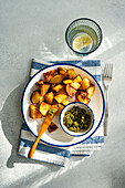 From above of crisp roasted potatoes beside a bowl of green pesto on a blue-trimmed plate, over a blue-striped napkin, with a glass of water with lemon slice in the background, on a textured surface in daylight