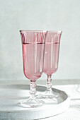 Ein Duo aus rosafarbenen Gläsern im Vintage-Stil steht auf einer spiegelnden Oberfläche, die in sanftes Sonnenlicht getaucht ist, vor einem minimalistischen Hintergrund. Generatives AI-Bild