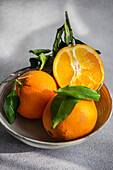 A pair of ripe oranges with lush green leaves sit in a simple bowl, casting shadows on a textured grey backdrop