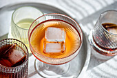 Draufsicht auf ein Sortiment strukturierter Gläser, die mit verschiedenen alkoholischen Getränken gefüllt sind, wobei ein markantes Glas mit einem großen Eiswürfel hervorgehoben wird