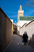 Ein Mann geht an einem klaren Tag eine enge Straße in der Medina von Fez entlang, mit dem Minarett im Hintergrund.