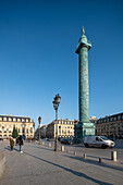Pedestrians walking near the Vendome Column under a clear blue sky.