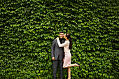 Frau küsst Mann auf die Wange vor einer grünen Hecke