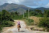 Cuba ,Trinidad,a man riding a horse walks on a deserted road in front of a landscape of green hills