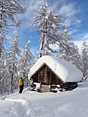 Österreich,Tirol,ein Mann mit einem gelben Rucksack steht in einem Lärchenwald vor einer frisch verschneiten Holzhütte