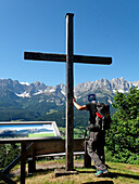Austria,Tyrol,Kitzbuhel,a man carrying a backpack is standing against a high wooden cross at Holenauerkreuz in front of the WILDERKAISER limestone range