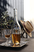 Flasche und Glas mit Getränk auf Tisch, Olivenzweig, Stuhl mit Strohhut im Hintergrund