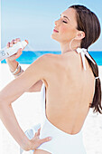 Young, dark-haired woman in white swimming costume spraying herself with sunscreen