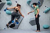 Young man teaching woman in her twenties how to climb on a climbing wall indoors