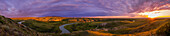 Ein Panorama der Landschaft und des Himmels bei Sonnenuntergang über dem Writing-on-Stone Provincial Park (Áísínai'pi) in Alberta, mit dem Milk River, der sich durch die Sandsteinfelsen windet, und den Sweetgrass Hills in der Ferne in Montana.