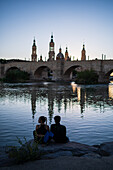 Junges Paar genießt den Sonnenuntergang am Ufer des Ebro, mit der Kathedrale-Basilika Unserer Lieben Frau von der Säule, Zaragoza, Spanien