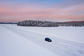 Luftaufnahme eines Autos, das auf einer vereisten Straße auf einem zugefrorenen See fährt, der von einem schneebedeckten Nadelwald umgeben ist, in der Abenddämmerung, Jokkmokk, Norrbotten, Schwedisch-Lappland, Schweden, Skandinavien, Europa