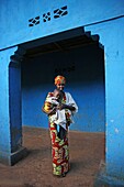 Rwanda, center of the country, woman in colorful loincloth with her baby in arms in front of the wall of a blue house