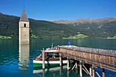 Italy, autonomous province of Bolzano, Lake Resia, boat moored to a pontoon with the steeple of an engulfed church emerging from lake waters surrounded by lush mountains