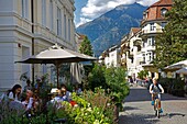 Italien, autonome Provinz Bozen, Meran, Kinder auf Fahrrädern und Menschen, die auf der Terrasse eines Cafés im Stadtzentrum sitzen, mit den grünen Bergen im Hintergrund