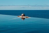 Spanien, Kanarische Inseln, La Palma, entspannte attraktive junge Frau beim Schwimmen im luxuriösen Infinity-Pool am Meer