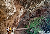 Spanien, Kanarische Inseln, La Palma, Touristen auf einem Wanderweg unter einem gewölbten Vulkangestein am Meer