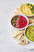 Top view assortment of classic, beet, and avocado hummus served with pita bread on a white plate, garnished with herbs and spices.