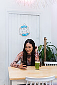 Positive female sitting at table with smoothie and browsing mobile phone while chilling at weekend in bar