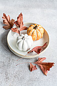 A serene autumnal scene featuring two pumpkins and dried brown leaves arranged on a simple ceramic plate, set against a textured gray background