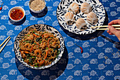 From above, a colorful display of Chinese cuisine with noodles and prawn dumplings on patterned plates, accented with condiments and chopsticks with an anonymous hand eating