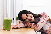 Positive female leaning on hand while sitting at counter with green smoothie and chilling in bar during weekend