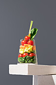 Fresh and colorful layered salad served in a transparent glass, elegantly displayed on a gray background. Healthy meal option featuring an array of vegetables like tomatoes, spinach, and carrots