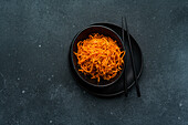 A top view of a black bowl filled with shredded carrot salad placed on a dark textured surface, accompanied by black chopsticks