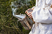 Crop anonymous beekeeper in protective suit standing in green garden with metal bee smoker