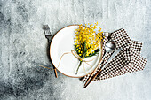 Top view of rustic-chic table setting featuring a delicate mimosa flower bouquet tied with twine, complemented by elegant cutlery and a checkered napkin