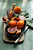 A vibrant display of blood oranges, some whole and others cut, on a dark plate, with a green cloth and citrus reamer in the background