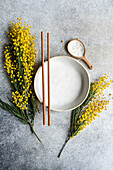 Top view of elegant minimalist table setting, with a ceramic plate and chopsticks, accented by the bright yellow of mimosa flowers against a textured grey background