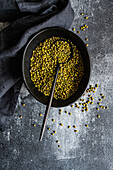 A rustic setting displays raw green mung beans scattered around and within a dark bowl, accompanied by a metal spoon on a textured, grey surface