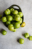 A bowl full of ripe organic green plums sits on a textured gray surface, with a few scattered plums around it These fruits are popular in various Mediterranean cuisines